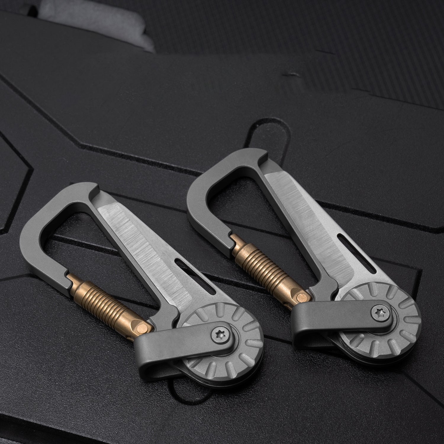 Two silver tactical multi-tool carabiners with bronze screws and rotating mechanisms, placed on a dark surface.